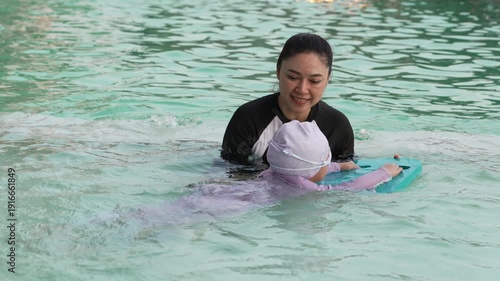 slow motion of preschool child girl learning to swim with board with her mother in swimming pool