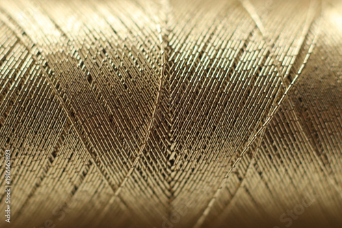 Macro close-up of a spool of golden thread showing detailed woven fibers and layered texture. The image highlights metallic sheen, craftsmanship, textile material, and abstract geometric patterns 
