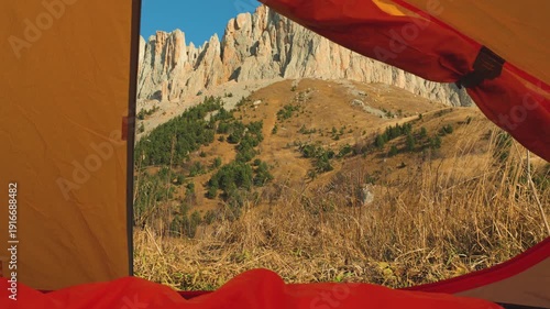 First-person view from inside a tent, with a red sleeping bag in the foreground. The open flap frames a sunny autumn mountain landscape with rocky cliffs. Cozy, adventurous mood