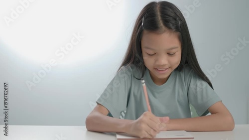 Young Asian girl concentrated on writing or drawing in a notebook. Concept of elementary education, homeschooling, creative thinking, and child development. Bright indoor setting with copy space.
