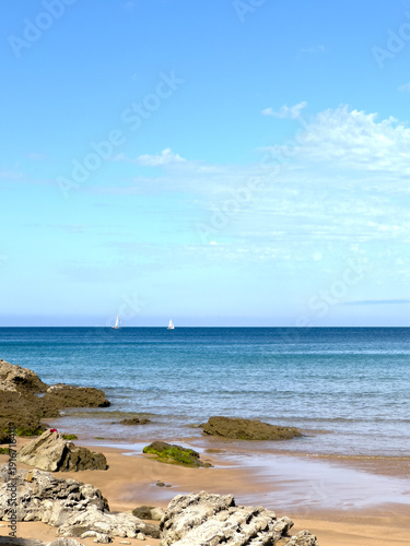 Two white sailboats on calm Atlantic horizon, rocky beach in foreground, subtle ocean and sky gradient from dark horizon to transparent blue shore, minimalist vertical seascape