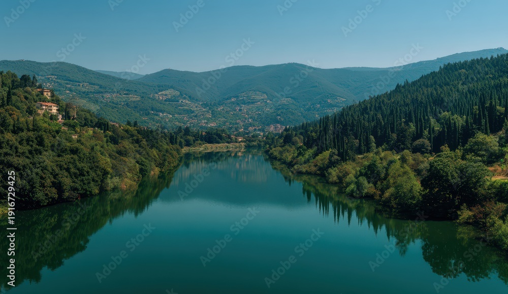 Naklejka premium Vibrant photo of Serene river winding through a dense green forest and rolling hills under a clear blue sky