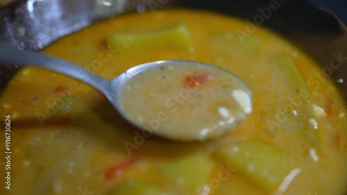 Extreme close-up macro shot of a silver spoon lifting a scoop of creamy, golden-yellow soup from a dark pot.