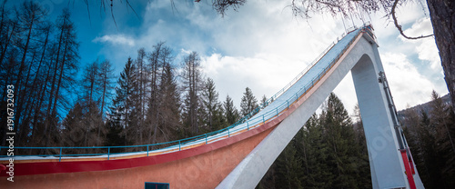 Foto Ski jump ramp rising above alpine forest in Cortina d Ampezzo, Italy, with dramatic concrete structure, blue sky, winter light, and towering pines creating a striking mountain sports scene