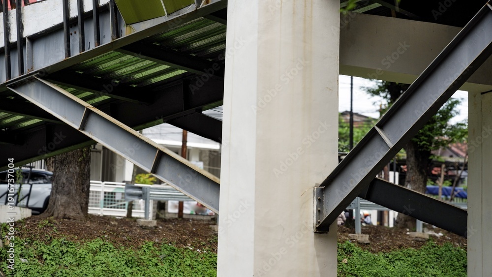 Fototapeta premium Close-up of steel beams and concrete pillars supporting a modern pedestrian overpass. An industrial architectural detail showing urban infrastructure and structural engineering.
