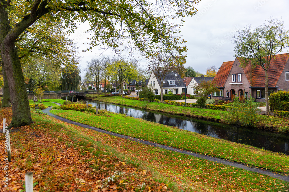 Fototapeta premium Hattem town Netherlands canal brick houses autumn foliage scenic idyllic landscape view. Quiet residential neighborhood with trees reflections pathway under cloudy sky peaceful travel atmosphere
