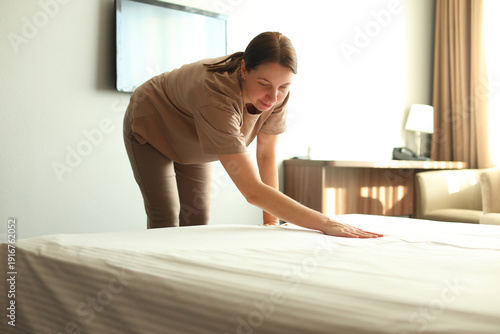 Housekeeper in brown uniform smoothing white bedsheet in bright hotel room, attentive, professional cleaning service in action.