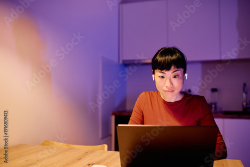Foto Adult woman with wireless earphones using laptop at home office during evening,
