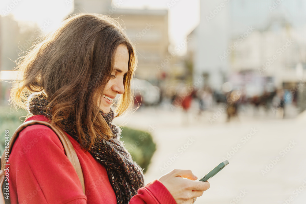 Fototapeta premium Smiling curly young woman in red sweater looking at smartphone screen on sunny city street.