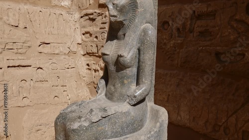 Ancient Egyptian goddess Sekhmet lion-headed statue in dark granite at Medinet Habu temple in Luxor Egypt, sacred deity sculpture with hieroglyphic inscribed walls background.