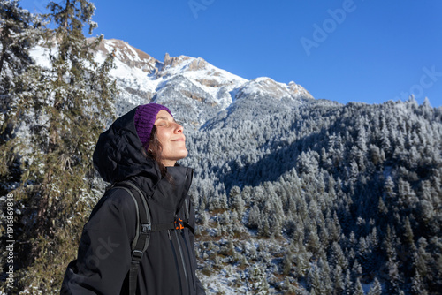 Profile of a woman with her eyes closed, breathing deeply and enjoying the sunlight in a snowy mountain forest. Peaceful expression with high peaks and frosted pine trees in the background. Concept of