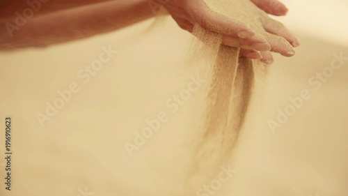 Cinematic close-up slow-motion shot of a hand letting fine, golden sand slip through fingers. The sand flows in a smooth, continuous stream, catching the warm sunlight. 