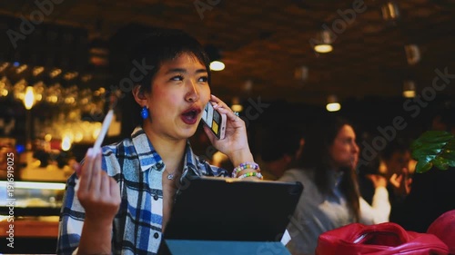 Serious Asian woman having mobile conversation in coworking space, digital nomad using smartphone and tablet for business, professional female expert phoning, career growth and technology