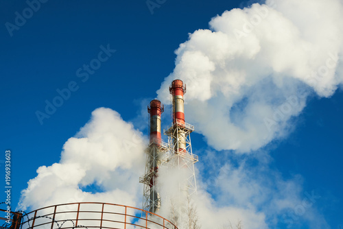 Two chimneys of a natural gas heating plant partially hidden by dense white steam. Water vapor and condensation cloud against a blue sky, winter utility background.