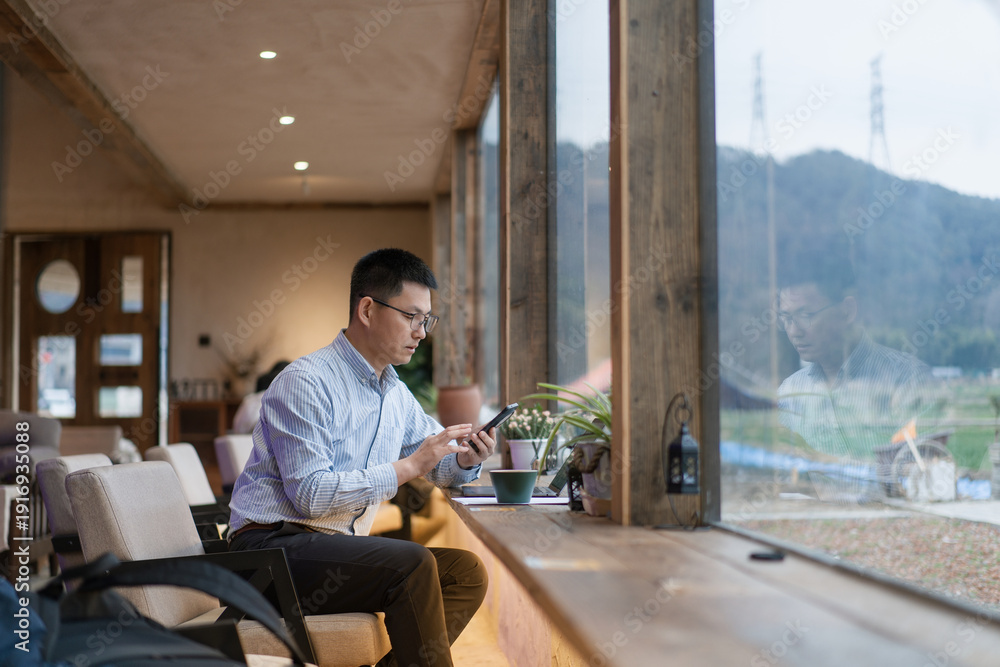 Fototapeta premium A young man wearing glasses uses his smartphone for work while sitting by the window in a modern wooden cafe with mountain views