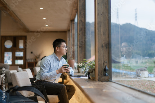 Fotografia A young man wearing glasses sits by the window holding a cup while gazing at the