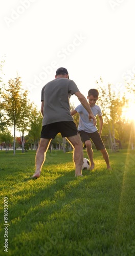 Vertical video of father and son playing soccer barefoot. Family plays soccer on green grass. Active dad and kid enjoy soccer training in summer park.