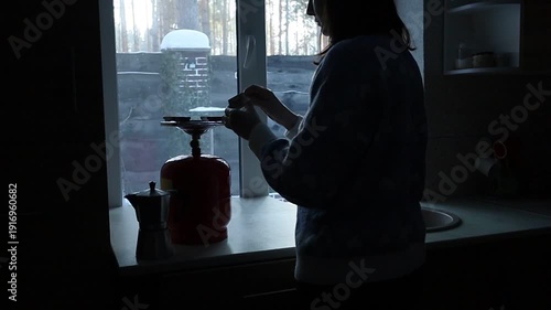 A young Ukrainian woman lights a gas stove with a match to make herself coffee during a power outage. The intimate kitchen scene highlights everyday resilience and adaptation to electricity shortages 