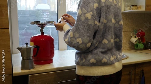 A young Ukrainian woman lights a gas stove with a match to make herself coffee during a power outage. The intimate kitchen scene highlights everyday resilience and adaptation to electricity shortages 