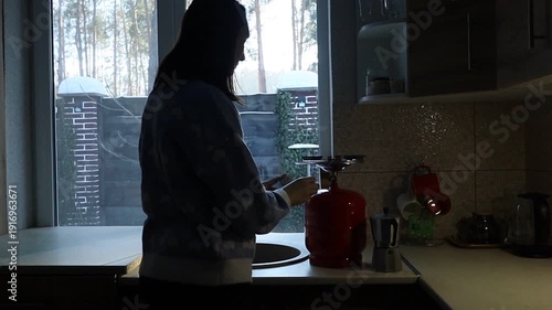 A young Ukrainian woman lights a gas stove with a match to make herself coffee during a power outage. The intimate kitchen scene highlights everyday resilience and adaptation to electricity shortages 