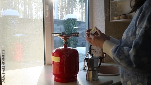 A young Ukrainian woman lights a gas stove with a match to make herself coffee during a power outage. The intimate kitchen scene highlights everyday resilience and adaptation to electricity shortages 