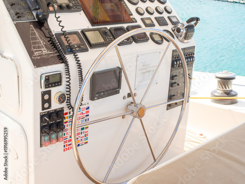 A detailed shot of a modern boat helm, showing a large stainless steel wheel, many gauges, a VHF radio, navigation screens, a set of flag stickers
