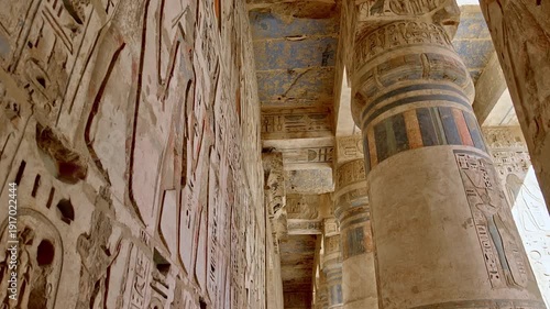 Medinet Habu temple colonnade with hieroglyphic inscriptions and relief carvings in Luxor Egypt, massive sandstone columns with papyrus capitals under dramatic blue sky.