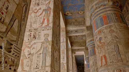 Medinet Habu temple colonnade with hieroglyphic inscriptions and relief carvings in Luxor Egypt, massive sandstone columns with papyrus capitals under dramatic blue sky. Mortuary Temple of Ramesses II