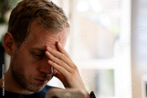 Young Caucasian man sits indoors during daylight, hand pressed to his forehead. He is experiencing stress, worry, and possibly a headache while looking down in deep thought