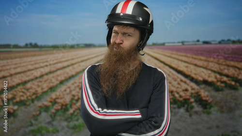 Foto Man with long beard wearing a helmet crossing arms and frowning in a tulip field under clear blue sky; defiance
