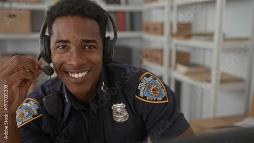 Foto Man police officer smiles and adjusts headset microphone while speaking at a desk in a building with shelves and boxes, badge visible; professional duty readiness