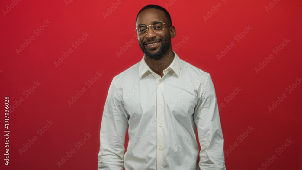 Obraz premium Man adjusting round eyeglasses with hands near face wearing white shirt in studio against red backdrop; confidence.