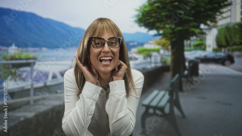 Woman in glasses and white shirt touching cheeks and laughing with tongue visible beside a bench on a street by a lake; playful joy.