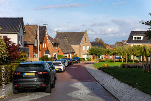 Cuadro en lienzo Dutch village street with brick houses, parked cars and curved roadway under blue sky with clouds