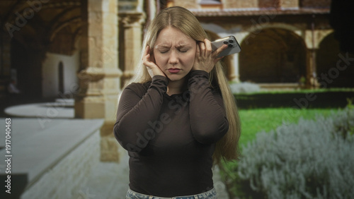 Tablou pe pânză Woman pressing hands to ears while holding smartphone to ear in cloister building; annoyance phone problem