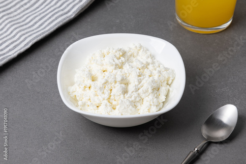 Cottage cheese in a bowl next to a glass of juice on a gray surface with a kitchen towel