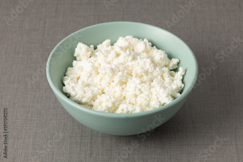 Fresh cottage cheese sits in a light blue bowl on a gray tablecloth during midday sunlight