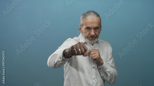 Man with hoary long hair and beard throwing punches with fists in studio blue backdrop wearing white shirt; determination grit.