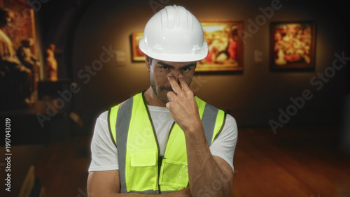 Fotografie Hispanic engineer man wearing white hardhat and reflective vest pinches nose against odor in building gallery; odor alertness