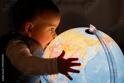 Valokuva A curious little boy, a few months old, looks at a glowing globe lamp in a dark room and touches the map with his hand, exploring the geography of the Earth, future generations