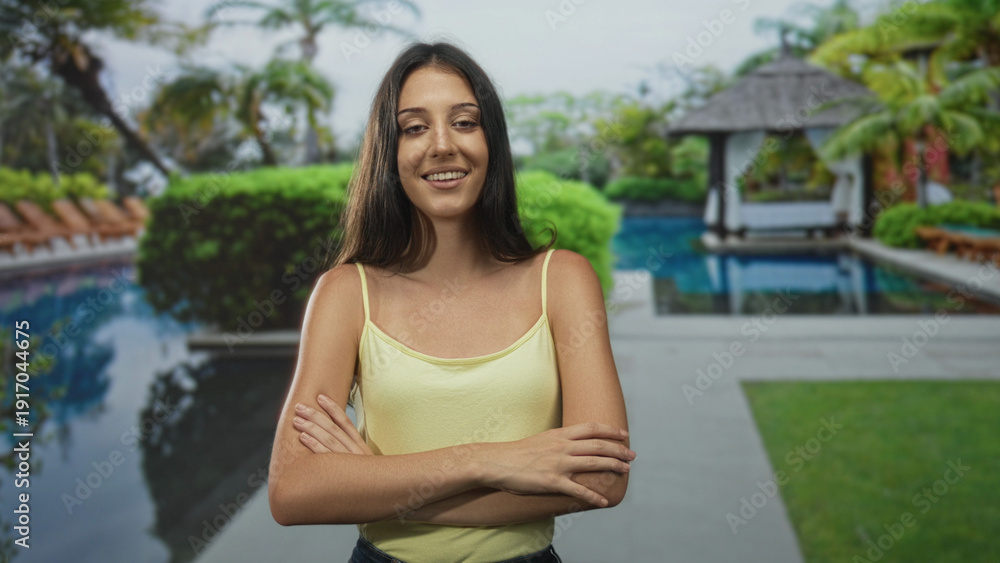 Naklejka premium Young hispanic woman with arms crossed in a yellow tanktop by hotel pool resort outdoors; confidence relaxation.