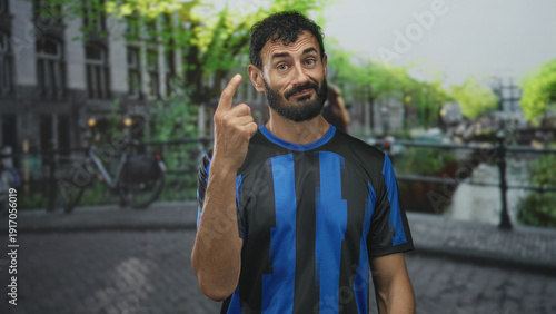 Cuadro en lienzo Man in soccer jersey points finger with a slight smirk standing on a street canal bridge beside bicycles and cobblestone; cheeky confidence