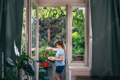 Young woman watering indoor plants on a balcony in a cozy apartment. Lush green urban garden visible through the open door. Sustainable lifestyle and mindful living at home
