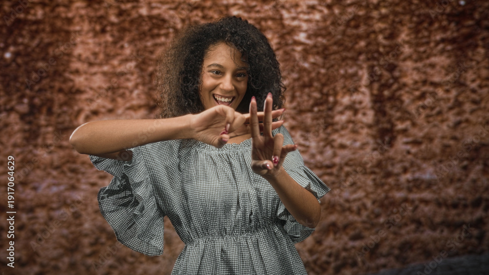 Obraz premium Woman forming a hashtag sign with her hands while smiling, wearing a gingham dress and curly hair against a textured backdrop in a studio setting; joy self expression.