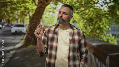 Man displaying a pinch gesture with one hand on a tree lined street near parked cars, wearing a plaid shirt and beard; quiet skepticism.