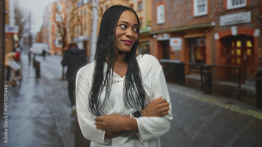 Fototapeta premium Black woman smiles with long braids and white blouse, pendant and watch visible while posing on a wet city street; confidence candid urban.