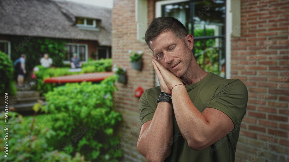 Fototapeta premium Man resting head on hands with hands pressed to cheek at a house building entrance, sleep gesture; calm rest.