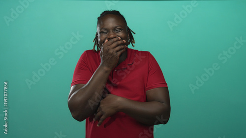 Man with hand covering mouth laughing in studio with teal backdrop and red polo shirt; joy playfulness.