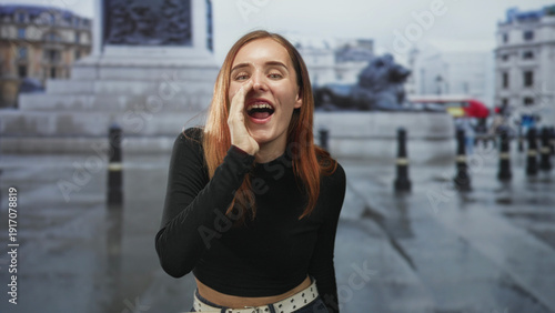 Redhead woman calls out with hand to mouth on wet street near building and statue, wearing casual jeans and studded belt; youthful joy.