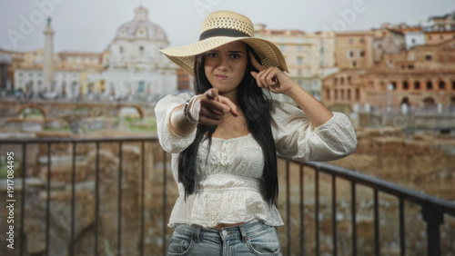 Schilderij op canvas Woman points finger to camera with straw sun hat and bracelets standing by railing in front of ancient building ruins; blame disapproval
